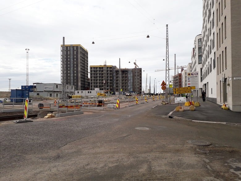 A large construction site. Completed buildings on the right-hand side of the street. Unfinished buildings behind them. A gritted street and construction site containers in the foreground.