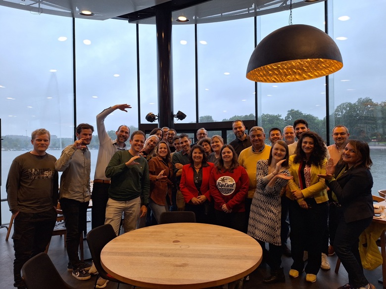 Suuri ryhmä ihmisiä seisoo ikkunan edessä sisätilassa, taustalla näkyy vesimaisema.