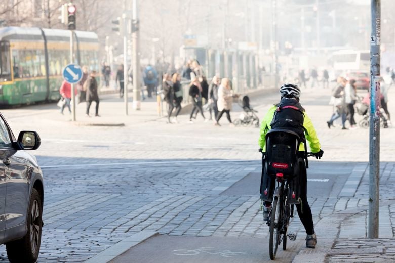 Pyöräilijä odottaa liikennevaloissa ja edessä kävelee jalankulkijoita sankassa katupölypilvessä.