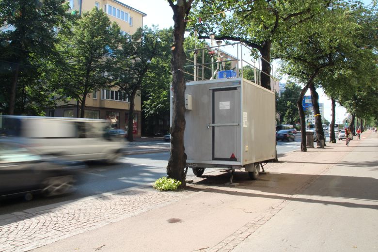 Ilmanlaadun mittausema seisoo pyörien päällä kadun varrella autojen ajessa ohi.