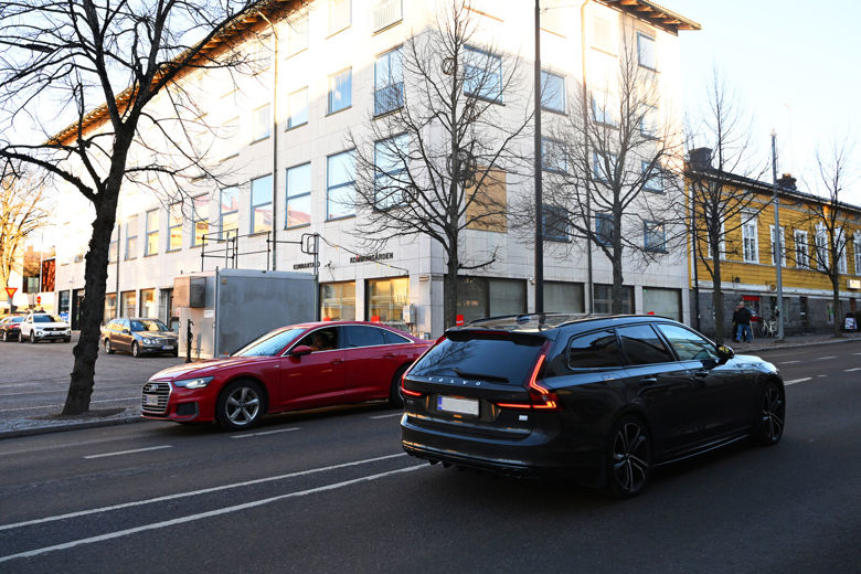 Kaksi autoa ohittaa kadun varressa olevan harmaan ilmanlaadun mittausaseman. Taustalla näkyy rakennuksia.