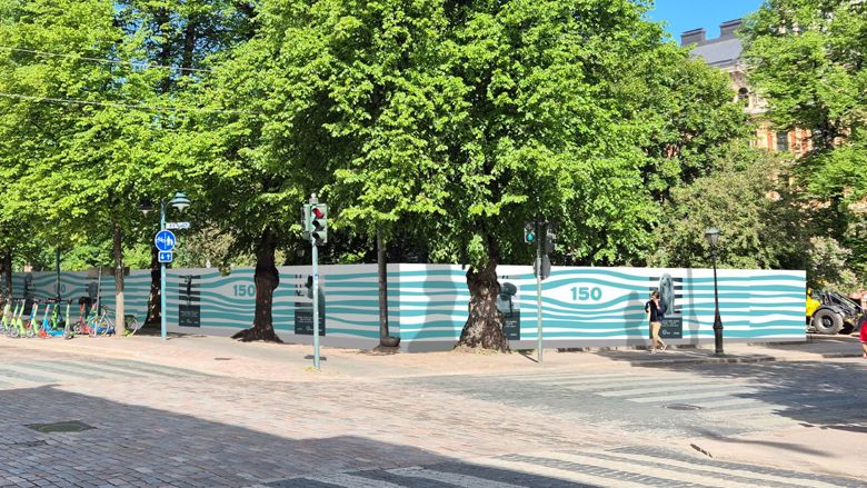 Havainnekuva Esplanadinpuiston työmaa-aidasta kaakkoiskulmalta katsoen. Työmaa-aidassa on HSY:n vesihuollon juhlavuoden ilme, eli virtaavan veden kuviota.