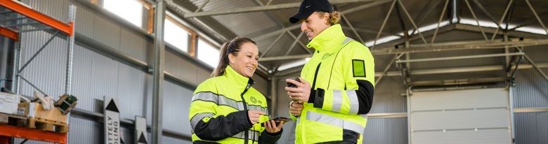 Två personer i höga siktningsvästar använder smartphones i ett industrilokaler.