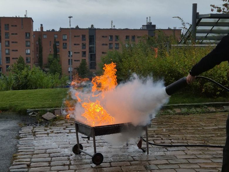 Henkilö sammuttaa palavaa tulta käyttäen käsisammutinta ulkona, taustalla kerrostalo ja puustoa.