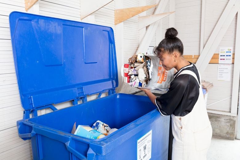 Nainen on jätetilassa laittamassa kartonkipakkauksia jäteastiaan.