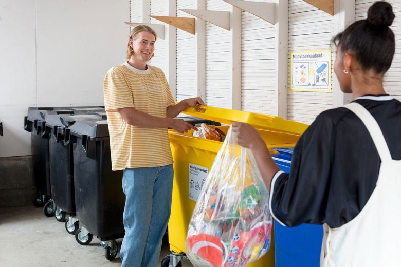 Kaksi ihmistä jätetilassa heittämässä roskiaan jäteastioihin. He hymyilevät toisilleen.