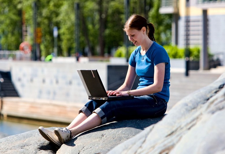 Hymyilevä nainen istuu kesällä aurinkoisella säällä kalliolla kannettava tietokone sylissään.