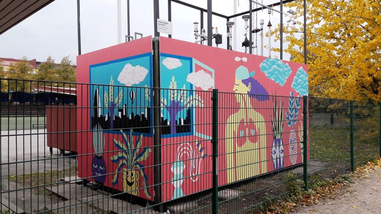 Brahe sports field air quality monitoring site in Kallio. Deciduous trees in autumn colours in the background. The measuring site is painted in bright colours, adorned with pictures of laughing plants, a window with floating clouds and a person whose throat and lungs have been painted to stand out.