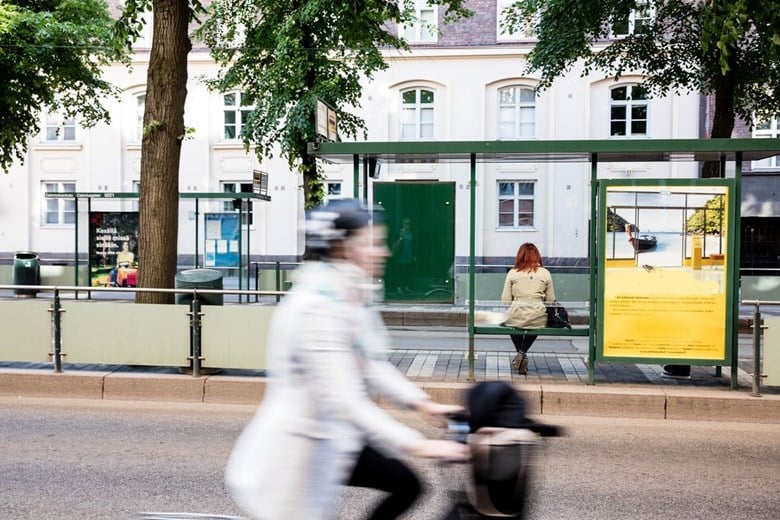 Cyclists travelling along the street so fast that they are slightly out of focus. Behind the bicyclist, a tram stop with a person waiting on the bench.  Trees on both sides of the stop. A row of buildings in the background.