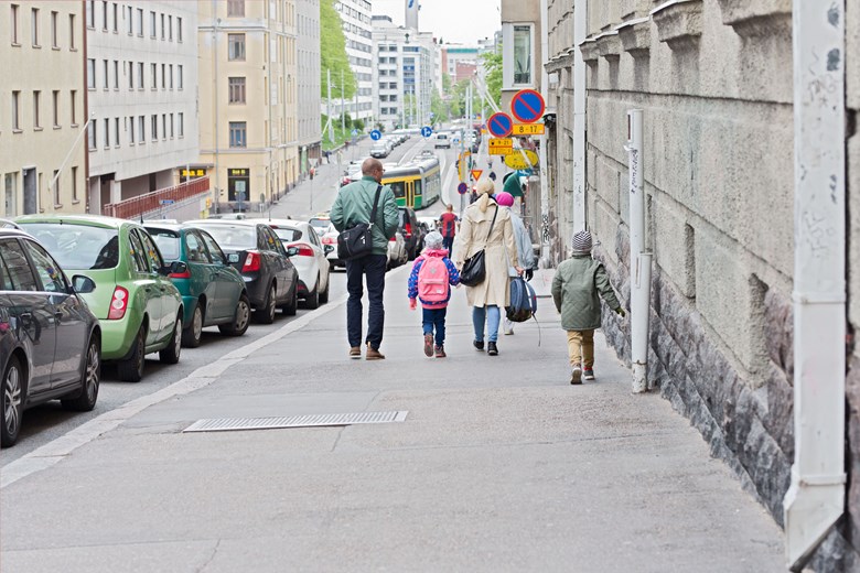Street sloping down, surrounded by buildings. There are cars parked along the street. There is also a tram in the background and there are a few trees in leaf by the street. On the pavement, there are adults and children walking with their backs facing the person looking at the image.