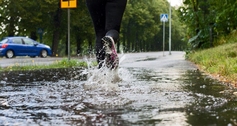 Kuvassa näkyy juoksevan lenkkeilijän jalat vettä tulvivalla kevyen liikenteen väylällä. Vesi roiskuu juoksukengistä. Tien vieressä kulkee auto.