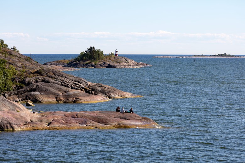Itämeri ja kallioista rantaa. Kuvassa näkyy kauempana kallioluoto, jossa on punainen pieni majakka. Etualalla näkyy kallioilla istuvia ihmisiä.